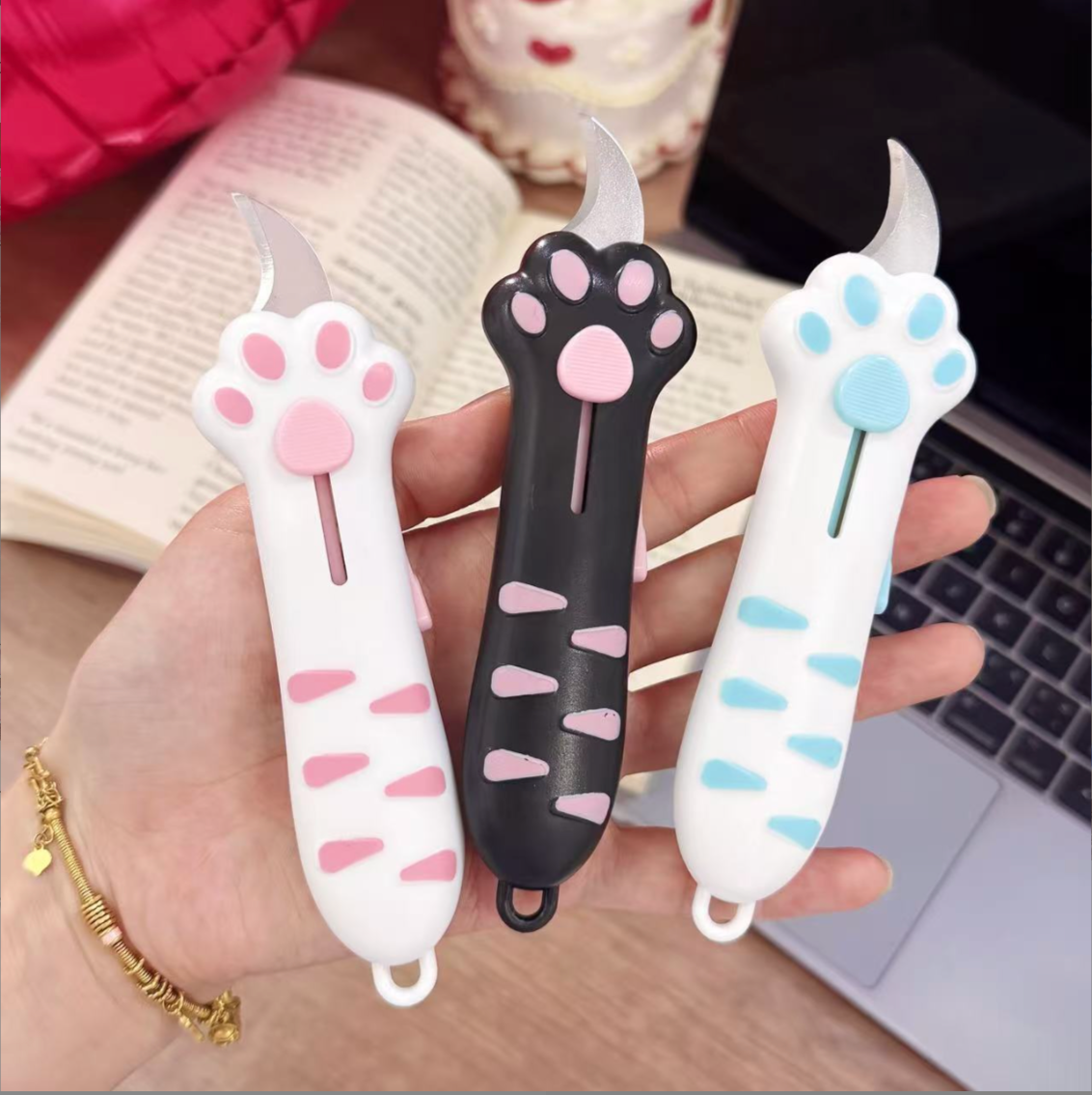 Hand holding three cat paw box cutters in White / Pink, Black / Pink and White / Blue.